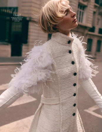 Fashion editorial shot of a woman in a textured white coat with feather epaulettes, outdoors with soft sunlight and blurred street background.