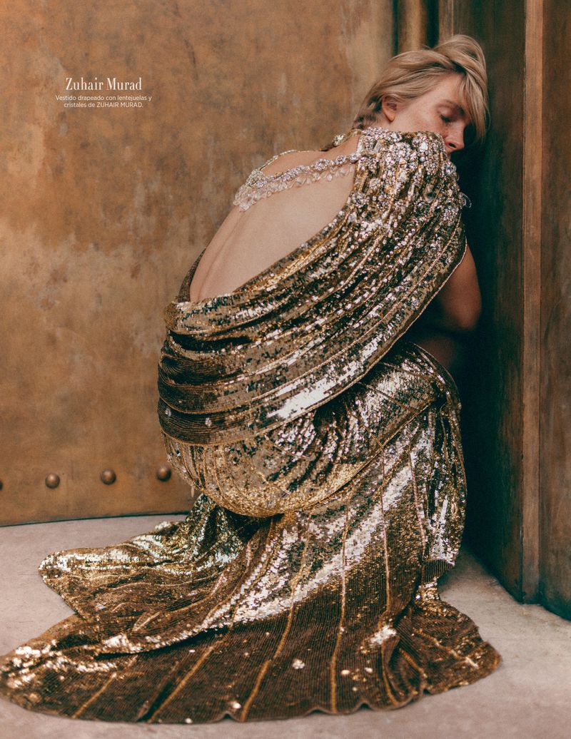 Blonde woman in a gold sequined gown with an open back, posing against a textured wall, showcasing high fashion and elegant design.
