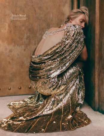 Blonde woman in a gold sequined gown with an open back, posing against a textured wall, showcasing high fashion and elegant design.