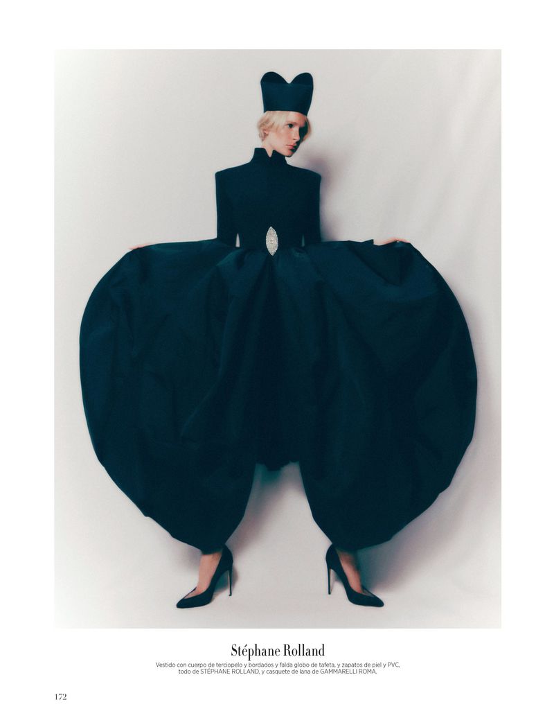 A slender adult female poses in an avant-garde black dress with voluminous trousers and a unique hat against a neutral background.