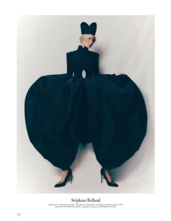 A slender adult female poses in an avant-garde black dress with voluminous trousers and a unique hat against a neutral background.