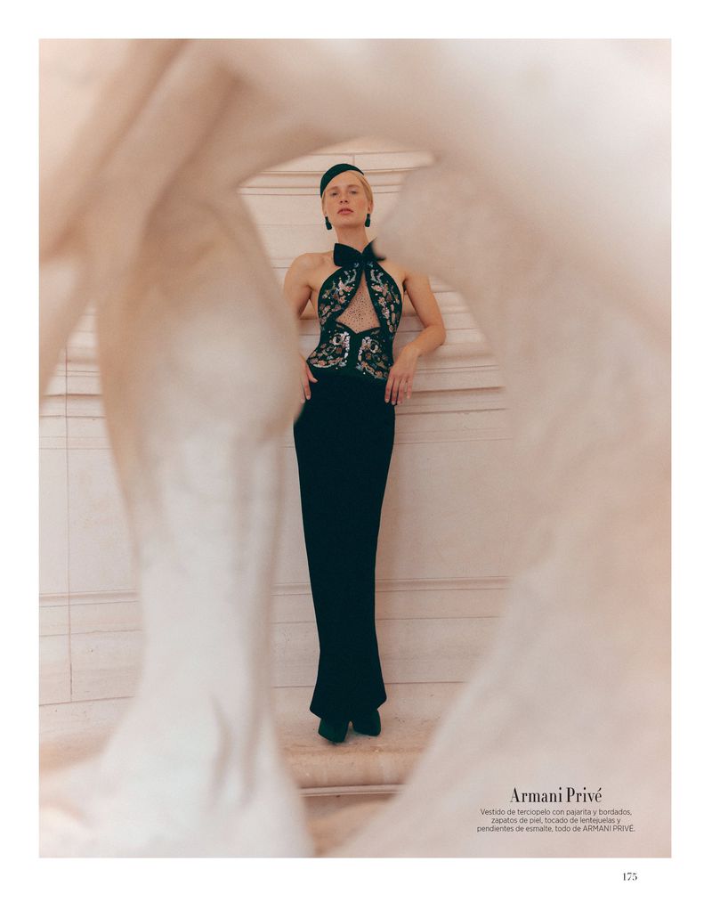 A woman in a black gown poses in front of a textured wall and blurred foreground, emanating elegance in this fashion photograph.