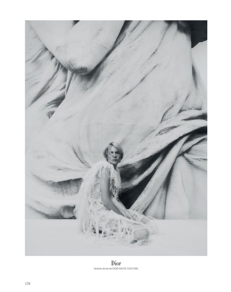 Monochrome fashion portrait of a woman in a light-colored, fringed dress sitting in front of an abstract, textured background. 