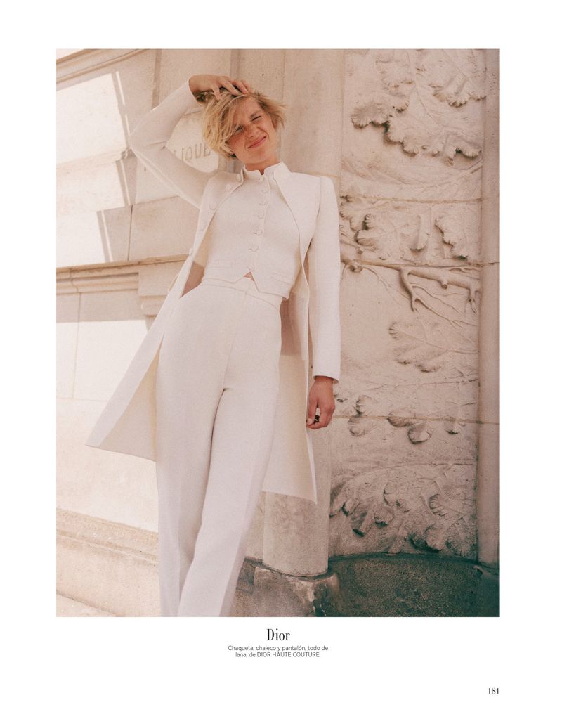 A woman in a white pantsuit poses in front of a stone wall with carved details in a high-fashion editorial shot for a magazine.