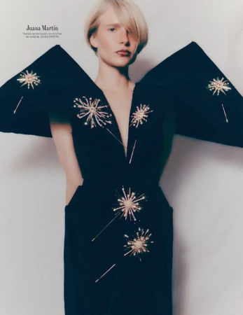 A woman with short blonde hair poses indoors wearing a black dress with starburst embellishments in a fashion editorial shot.