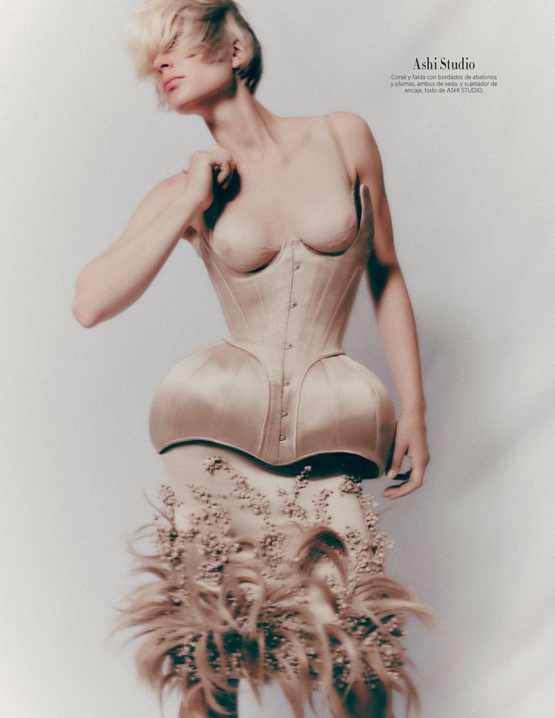 Fashionable woman in corset dress poses in studio, showcasing unique design with feathered detailing and blonde hair.