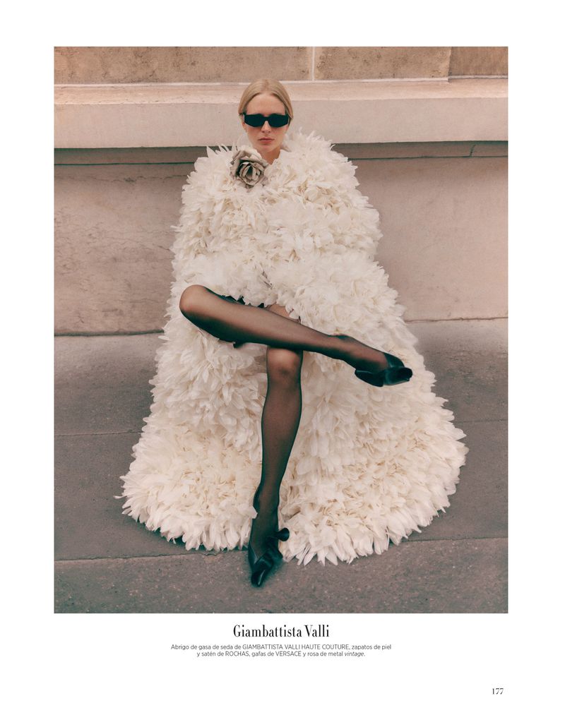 Fashion editorial shot of a fair-skinned adult woman in a feathered cape, sunglasses, and black tights, sitting against a stone wall.