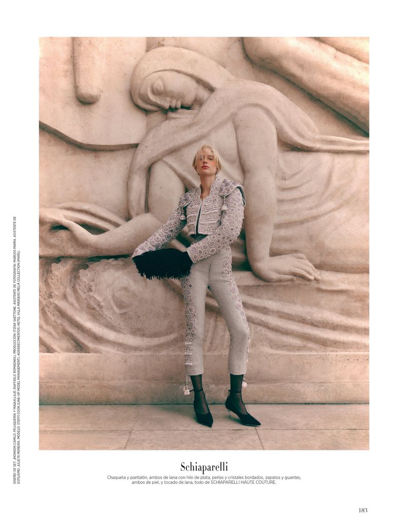 A fair-skinned fashion model in an embellished white suit with black accessories posing in front of a stone relief sculpture.