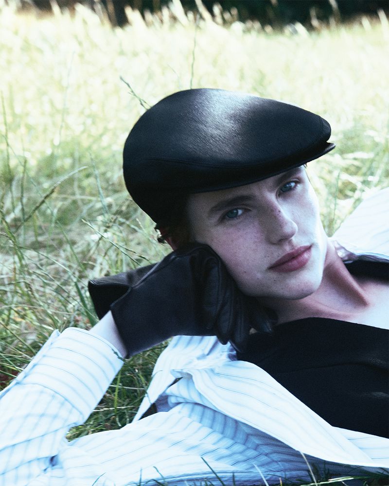 Portrait of a woman with fair skin and blue eyes wearing a black driving cap, black top, and striped shirt laying in the grass.