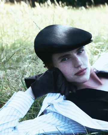 Portrait of a woman with fair skin and blue eyes wearing a black driving cap, black top, and striped shirt laying in the grass.