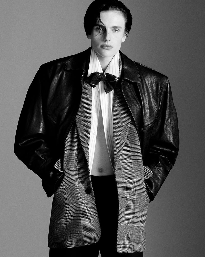 Monochrome fashion shot of a model in an oversized leather jacket, shirt, and blazer with a bow tie against a grey background.
