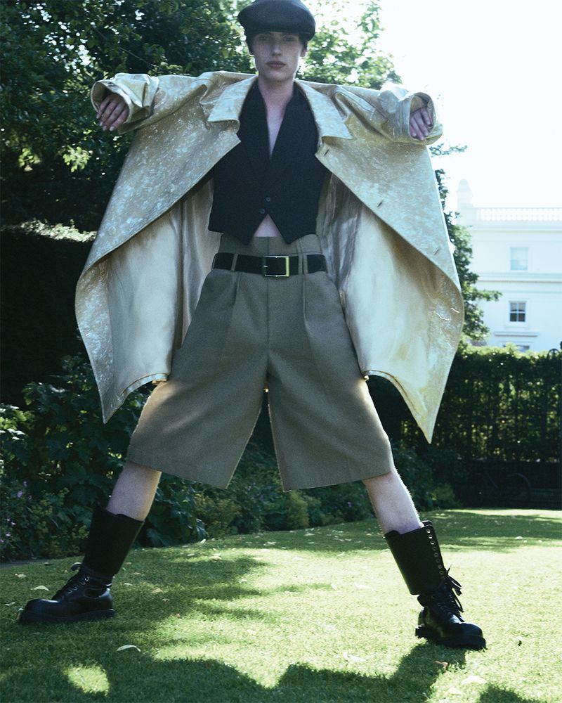 Person in a gold coat, black top, and brown shorts strikes a pose outdoors on a lawn with a house and trees in the background.