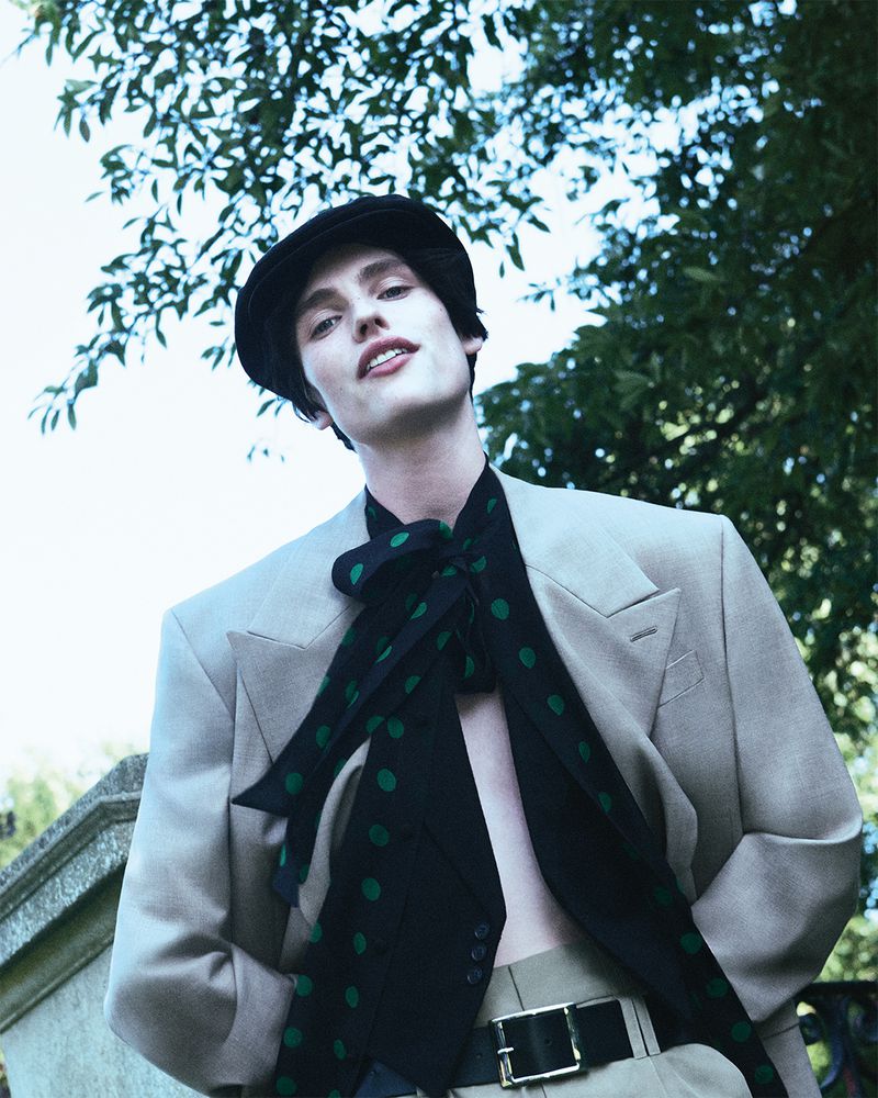 Portrait of a light-skinned male model in an unbuttoned tan suit, black polka dot scarf, and cap posing outdoors with green trees overhead.
