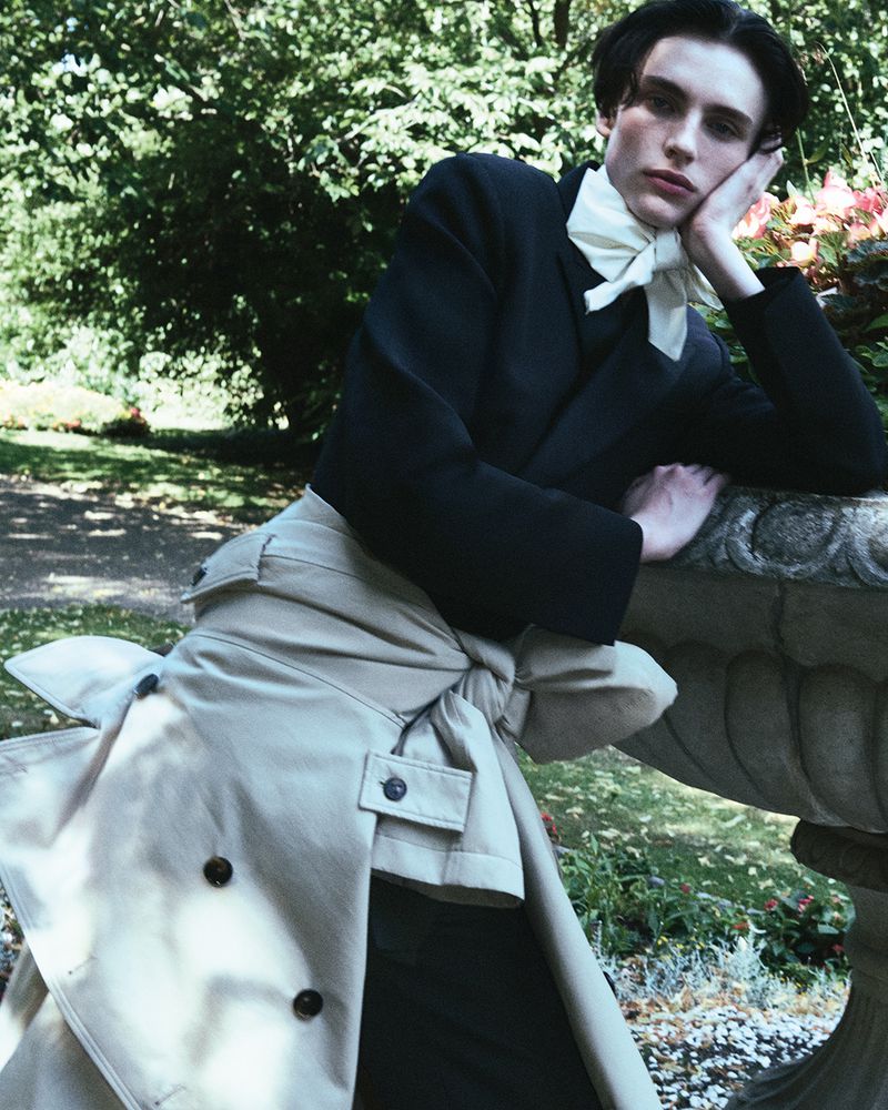 Young adult male fashion model poses in a garden wearing a black cropped jacket, white tie, beige trousers, and a trench coat.