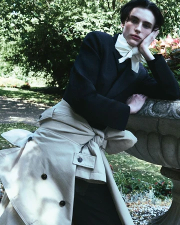 Young adult male fashion model poses in a garden wearing a black cropped jacket, white tie, beige trousers, and a trench coat.