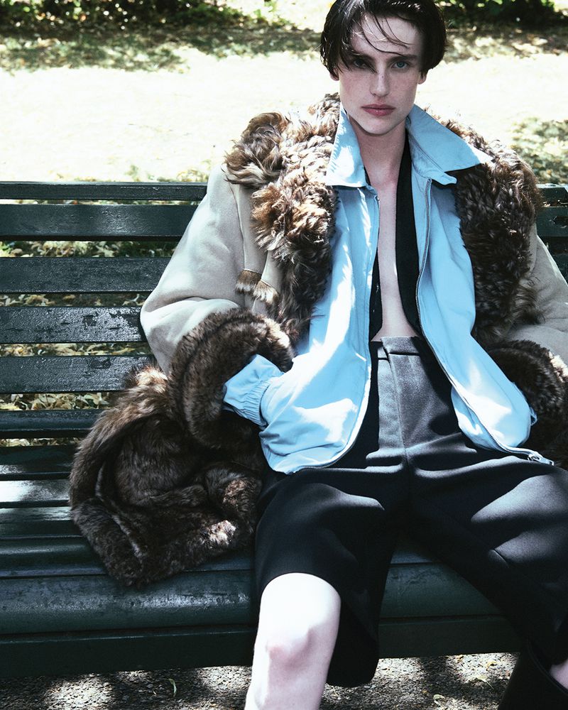 A young adult model is sitting on a park bench wearing a light blue jacket, dark trousers, and a light coat with fur trim.