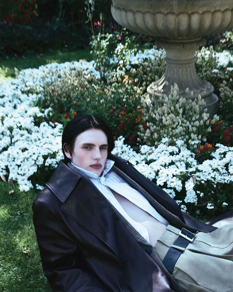 A young adult lies on green grass wearing an unbuttoned white shirt, a brown leather coat, and light-tan pants with a black belt. 