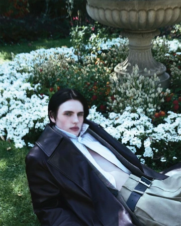 A young adult lies on green grass wearing an unbuttoned white shirt, a brown leather coat, and light-tan pants with a black belt. 