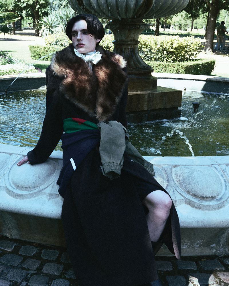 Fashionable adult in overcoat poses by marble fountain with greenery in background, photographed at eye-level, medium shot in natural light.