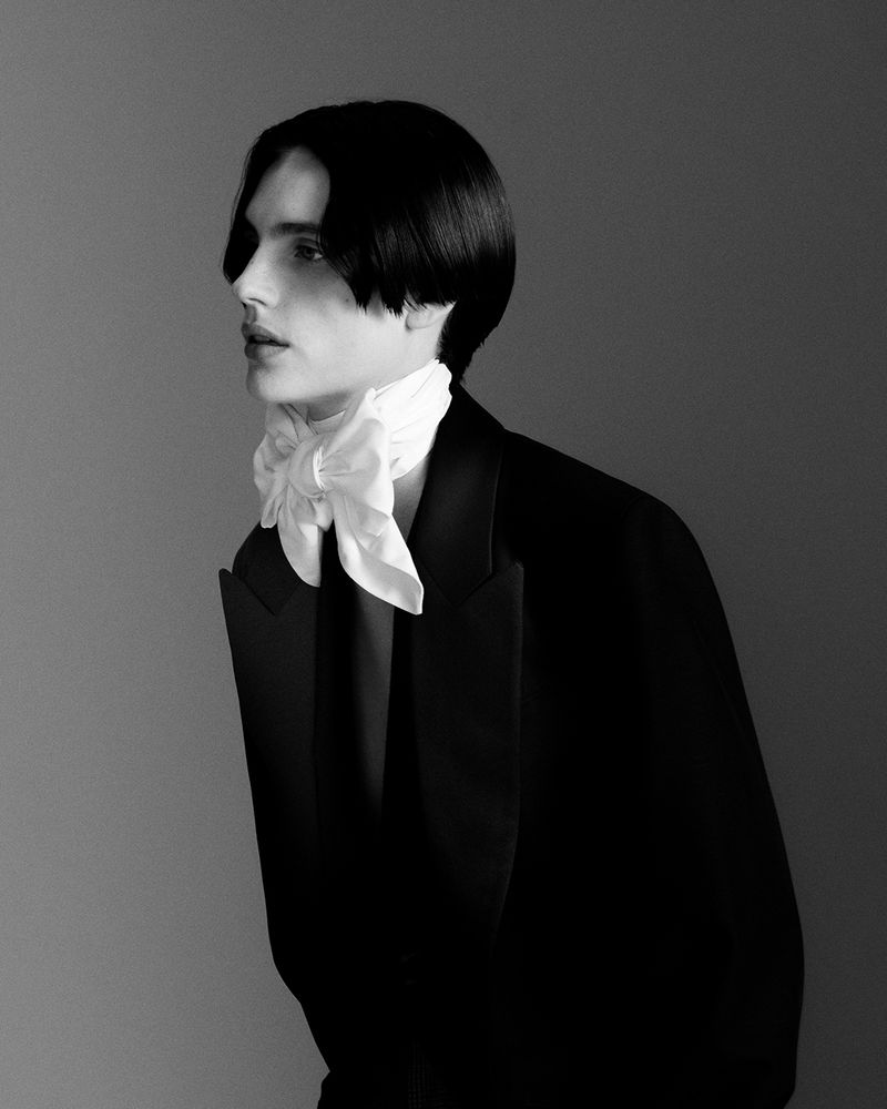 Black and white portrait of a young adult man with dark hair, wearing a black suit jacket and white tied scarf, looking left.