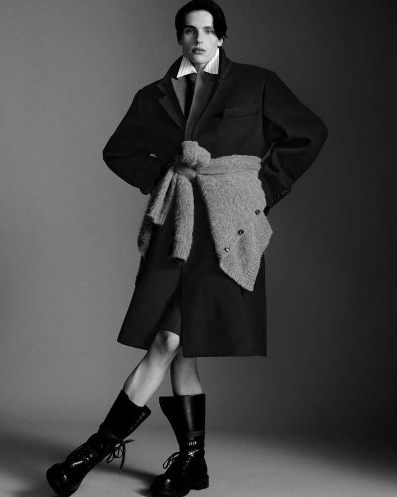 Black and white fashion shot of a slender young man in a dark coat, boots, and sweater, posing in a photo studio with a neutral background.