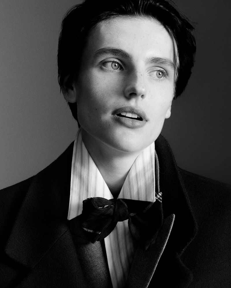 Black and white portrait of an adult with dark hair, wearing a bow tie and collared shirt with a coat, looking up and to the side.