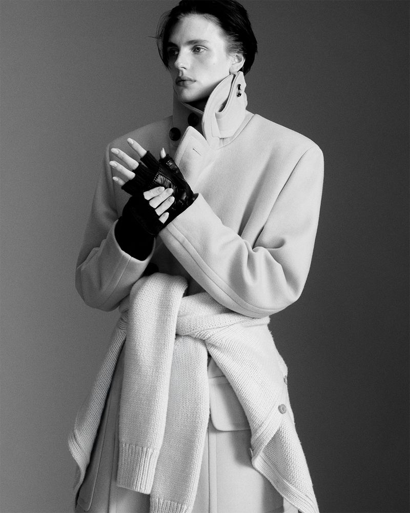 Monochrome fashion shot of a slender person posing in a light-colored coat, fingerless gloves, and sweater tied around the waist in a grey studio.