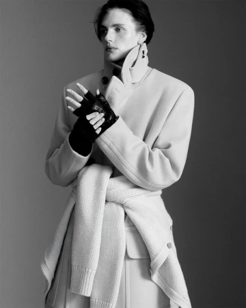 Monochrome fashion shot of a slender person posing in a light-colored coat, fingerless gloves, and sweater tied around the waist in a grey studio.