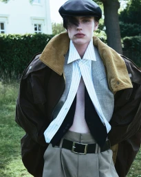 Model wearing a flat cap, oversized brown coat, striped shirt, and waistcoat standing outdoors in front of a building.