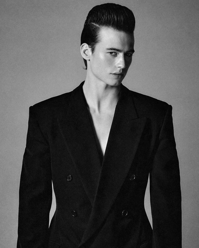 Black and white portrait of an adult male with pompadour hairstyle, wearing a double-breasted blazer, set against a neutral backdrop.