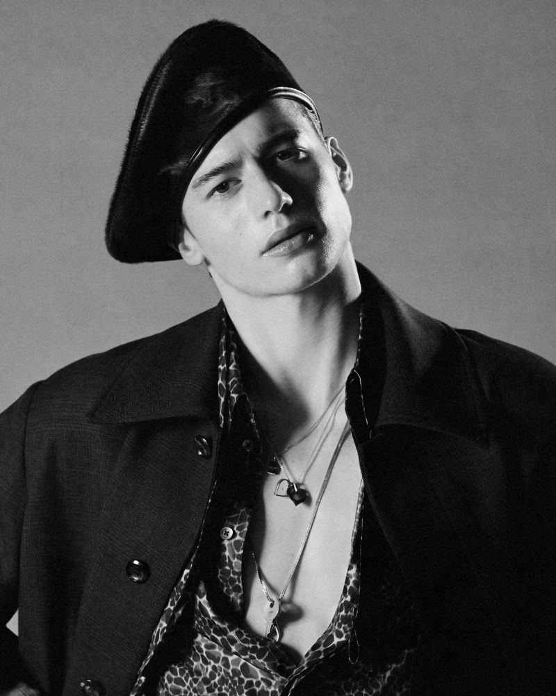 Black and white portrait of a young adult male wearing a beret, patterned shirt, jacket and necklaces in a studio setting.