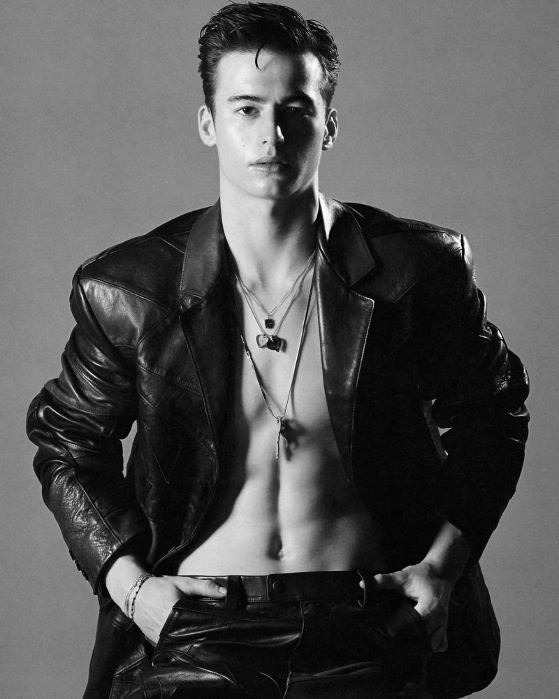 Black and white portrait of a young male fashion model wearing a leather jacket and pants with multiple pendant necklaces.