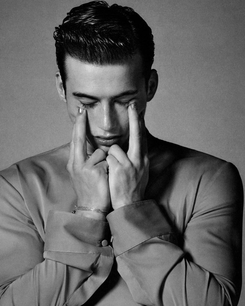 Striking black and white portrait of a young adult male with dark hair, eyes closed, and hands touching face in a moment of introspection.
