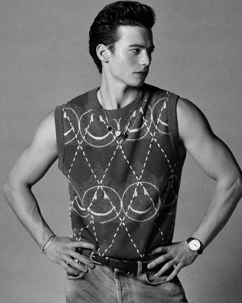 Black and white portrait of a young adult male in a patterned vest with arms on his hips looking off to the side.