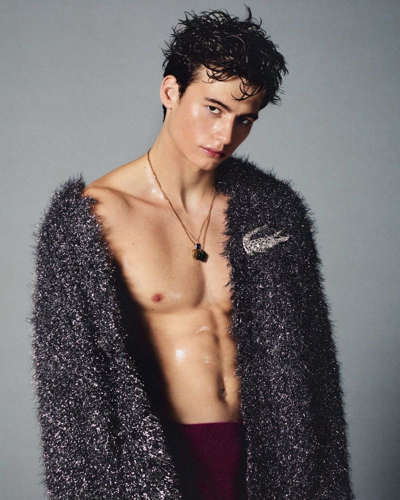 Attractive young man with bare chest poses wearing a fuzzy silver jacket and a necklace, indoors, with smooth skin and dark hair.