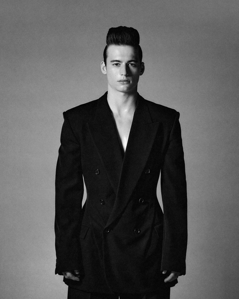 Monochrome portrait of a stylish young adult male with an elegant pompadour hairstyle, wearing a double-breasted suit jacket against a grey background.