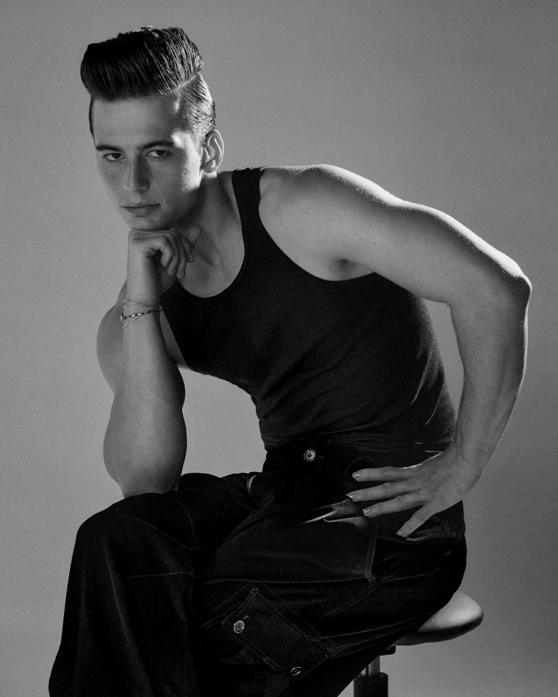 Monochrome portrait of a young man with a pompadour hairstyle, wearing a tank top and baggy pants, seated on a stool against a neutral backdrop.