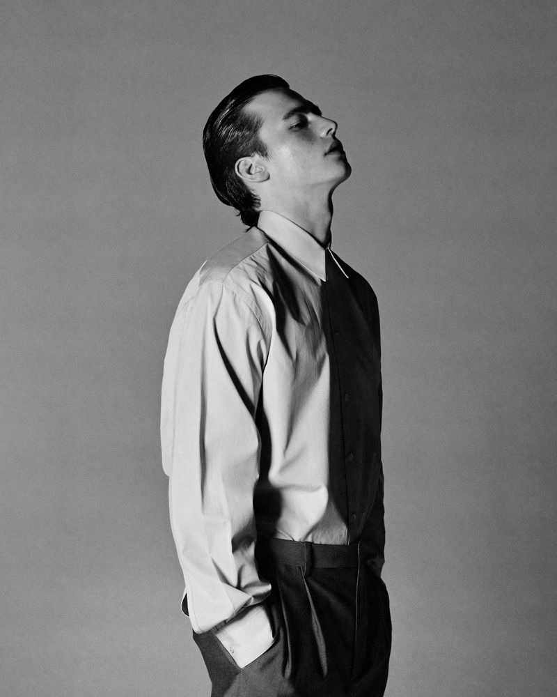 Monochrome portrait of a young adult man in dress shirt and dark pants, looking up with head tilted back against gray studio backdrop.