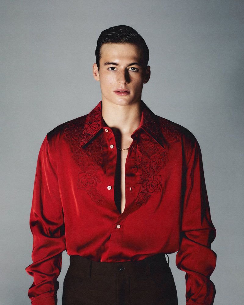 Portrait of a young adult man in a red satin shirt with a paisley design and black pants posing in a photo studio with a gray background.