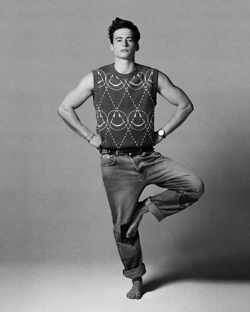 Black and white studio shot of a young adult man standing in a yoga tree pose wearing a sweater vest and jeans, arms on hips, looking at camera.