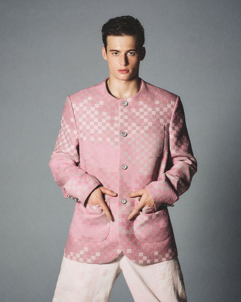 Male fashion model in a pink checkered jacket and white pants posing in a photo studio against a gray background in a medium shot.