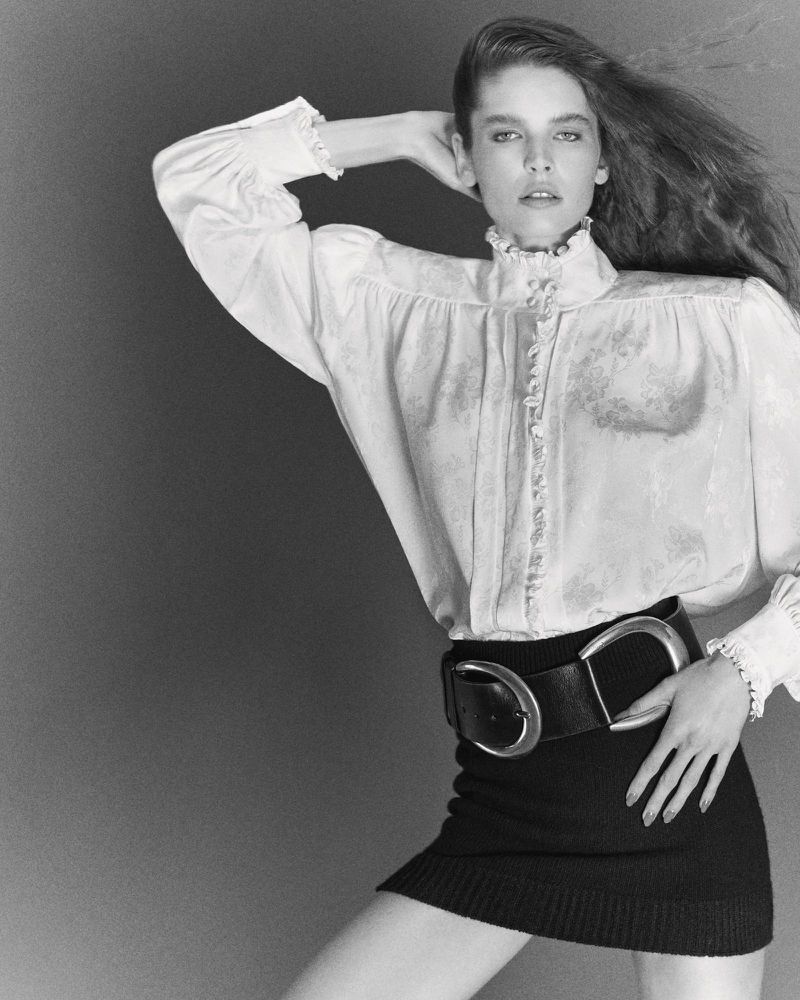 Monochrome portrait of a woman with long hair wearing a white blouse, black skirt, and a large belt against a neutral background.
