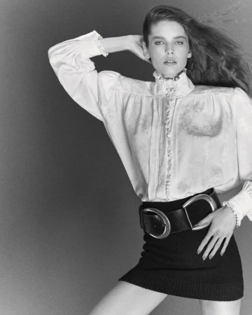 Monochrome portrait of a woman with long hair wearing a white blouse, black skirt, and a large belt against a neutral background.