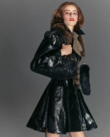 Fashionable woman in a shiny black coat and skirt holding a black leather purse against a neutral grey background in a studio shot.