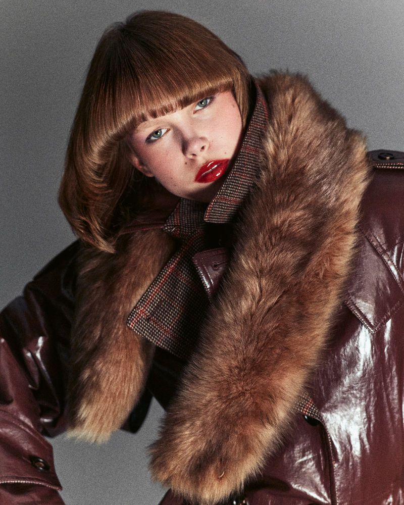 Close-up of a person with a stylish bowl cut and red lipstick, posing in a shiny brown coat with a large fur collar.