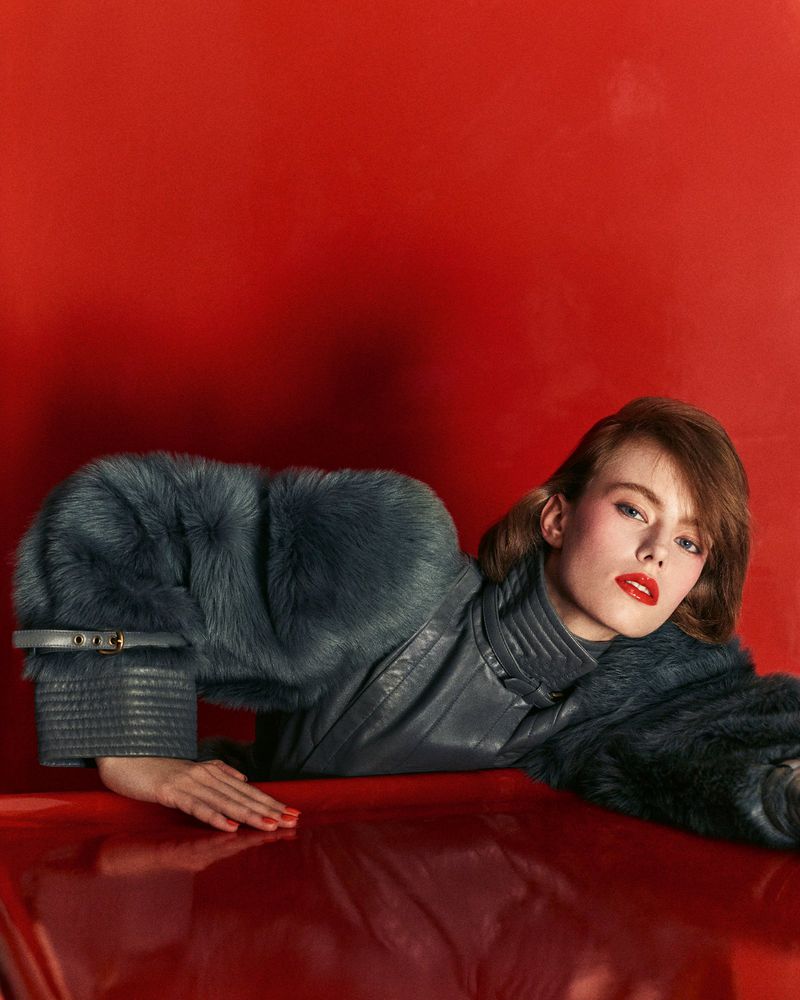 Glamour shot of a woman in a grey fur-trimmed jacket with red lipstick, posing against a glossy red surface and a red background.