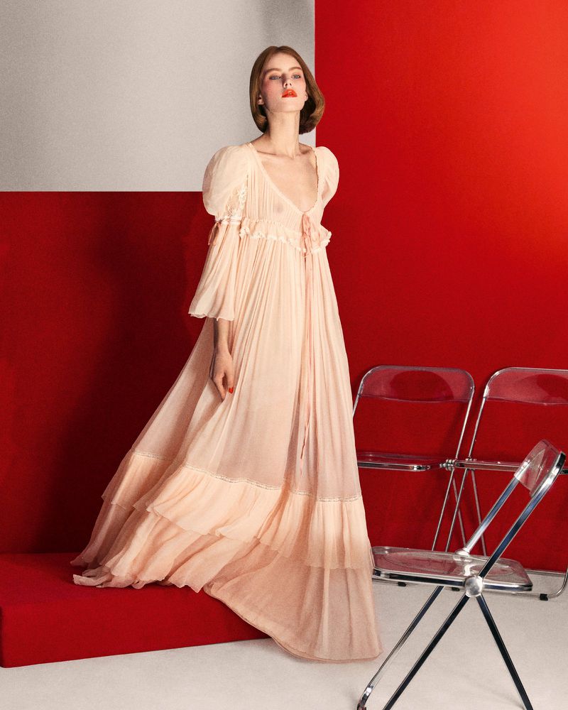 A redhead woman is posing in a long, light-peach dress with layered ruffles next to transparent acrylic chairs on a set with a white-red background.