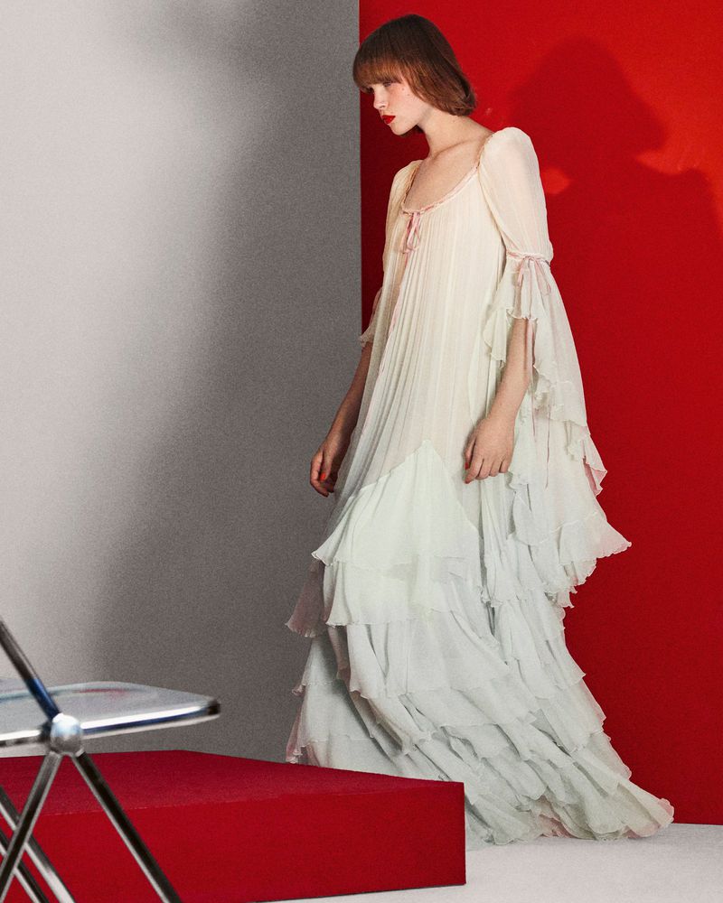 Elegant woman in a layered white chiffon dress poses against a vibrant red and grey wall in a fashion photo studio setting.