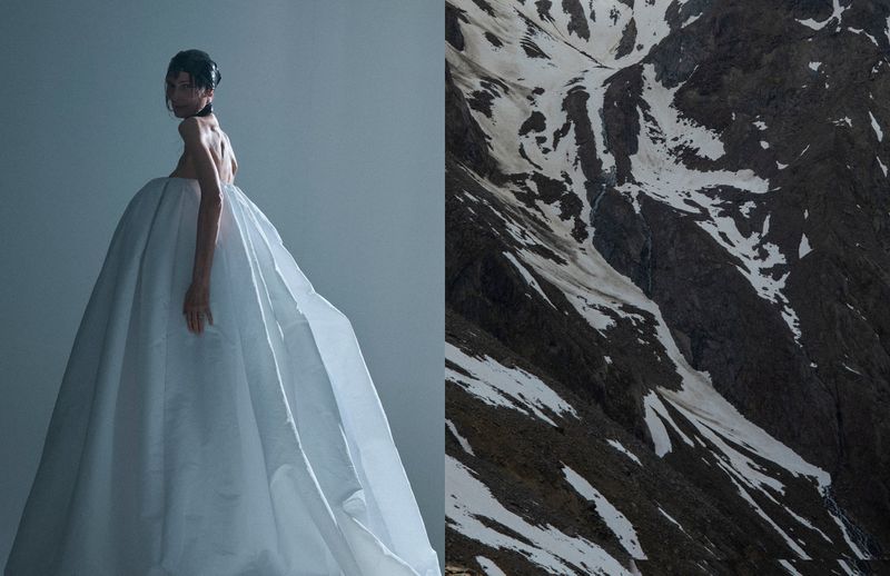 Woman in oversized white dress poses against a mountain backdrop. The dress is a dominant feature, contrasted by the dark, rocky terrain.