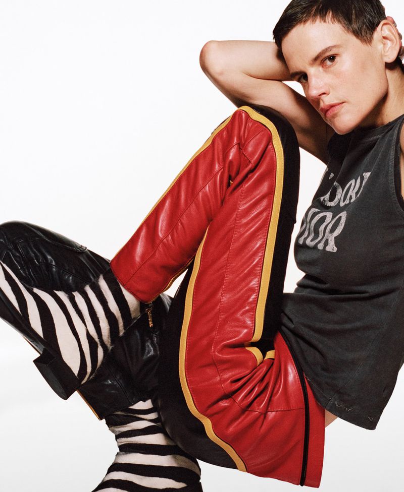 Adult woman with short hair poses in a tank top, red and yellow leather pants, and zebra-striped boots against a white backdrop.